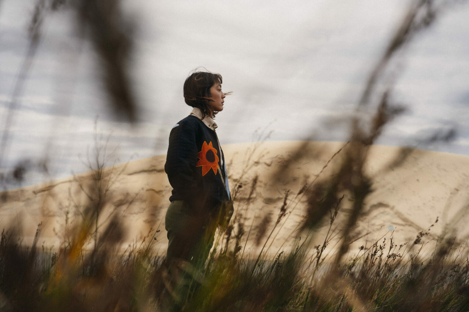 Personne portant le Carbon Black Kigo Zipped Sweatshirt de la marque OLOW dans un paysage naturel de dunes et d’herbes sauvages, vêtement au design graphique et éthique. Person wearing the OLOW Carbon Black Kigo Zipped Sweatshirt in a natural landscape of dunes and wild grass, featuring a graphic, ethical design.